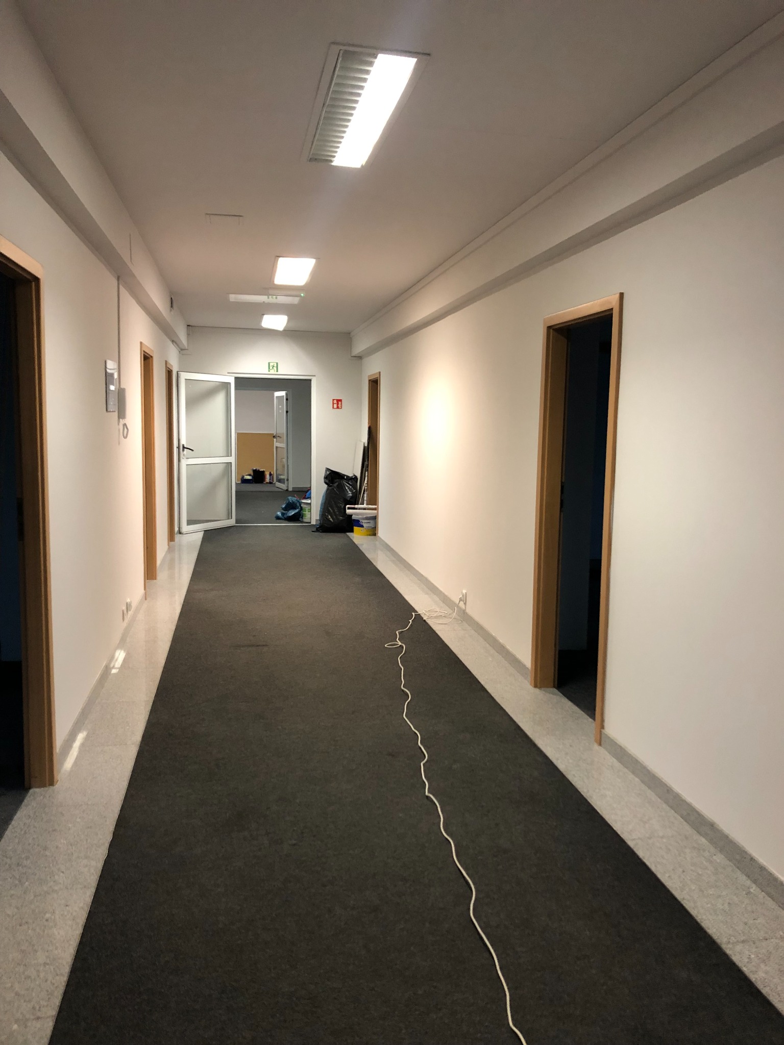 Długi korytarz biurowy z nowo pomalowanymi białymi ścianami, widoczne drzwi z drewnianymi ościeżnicami i ciemna wykładzina na podłodze, kabel elektryczny ciągnący się wzdłuż ściany.