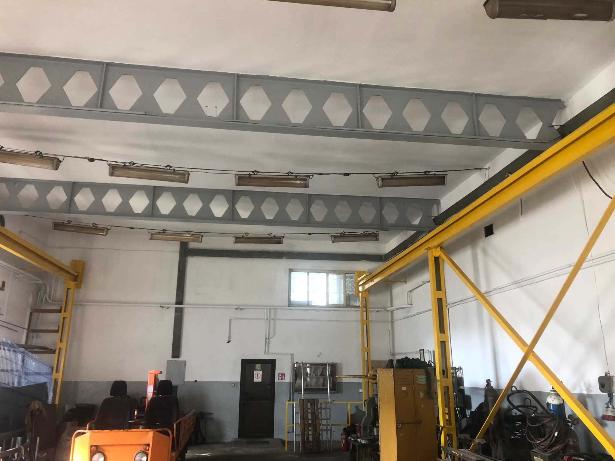 Wnętrze hali przemysłowej z szarymi, pomalowanymi belkami stropowymi o strukturze plastra miodu, żółtą suwnicą, oświetleniem i sprzętem warsztatowym.