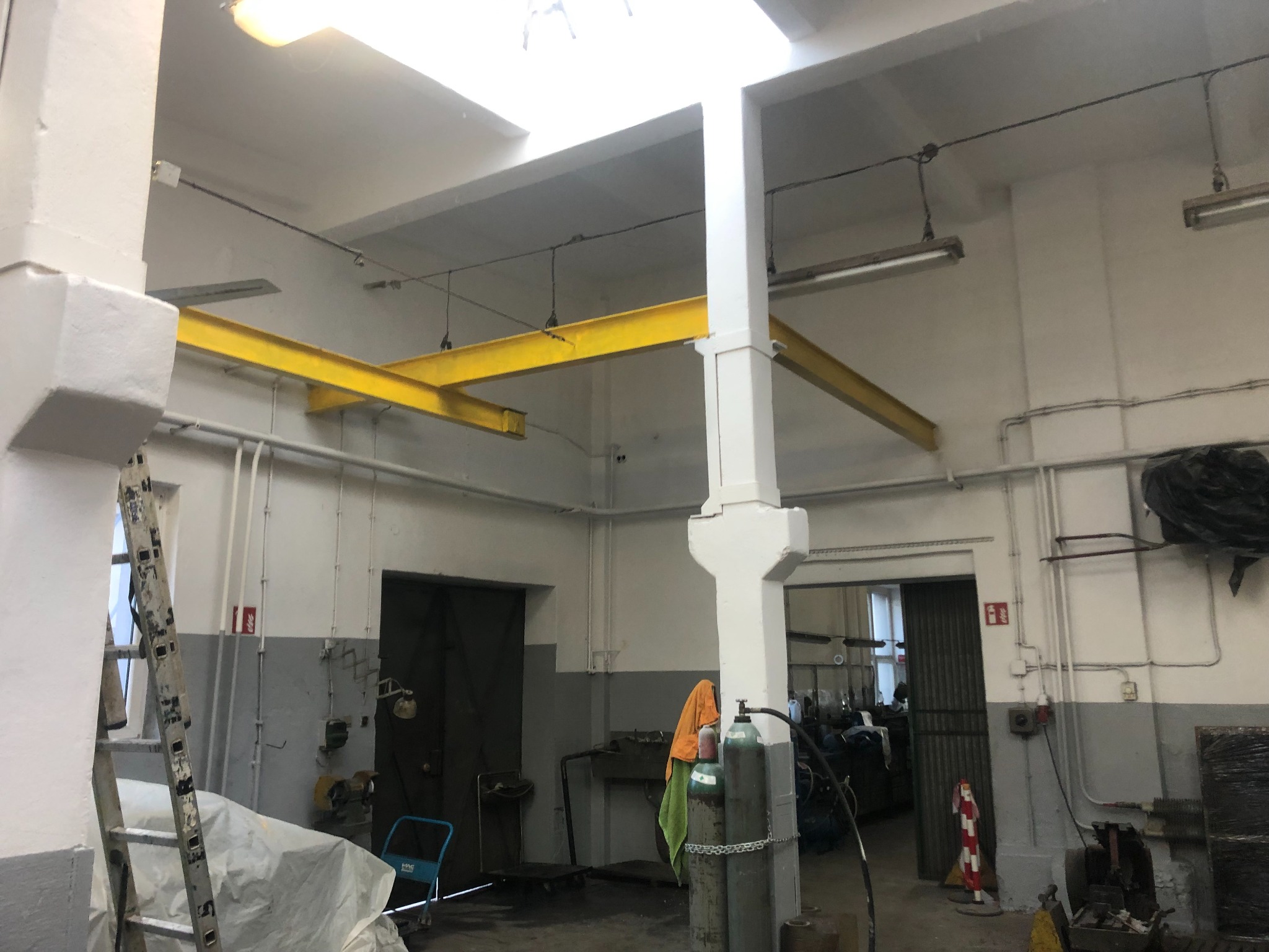 Wnętrze hali produkcyjnej po odświeżeniu, widoczne pomalowane na biało ściany, kolumny i sufit, z żółtymi belkami konstrukcyjnymi pod sufitem, drabina oparta o ścianę, butle z gazem i metalowe...