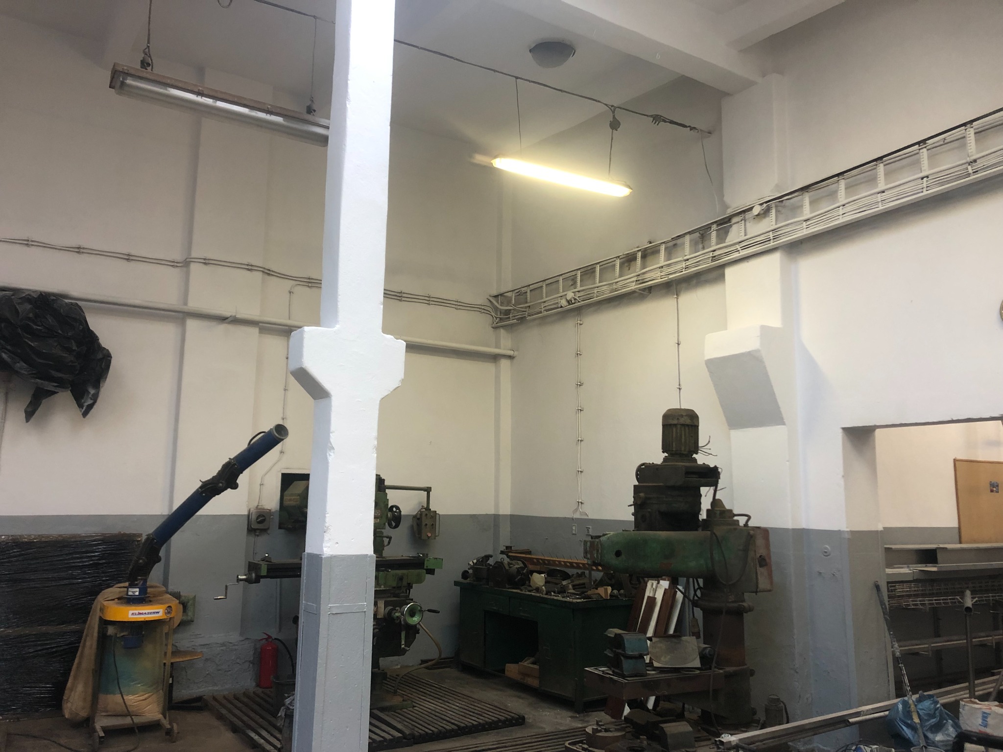 Wnętrze warsztatu z maszynami do obróbki metalu, kolumna pomalowana na biało-szaro, widoczne instalacje elektryczne pod sufitem.