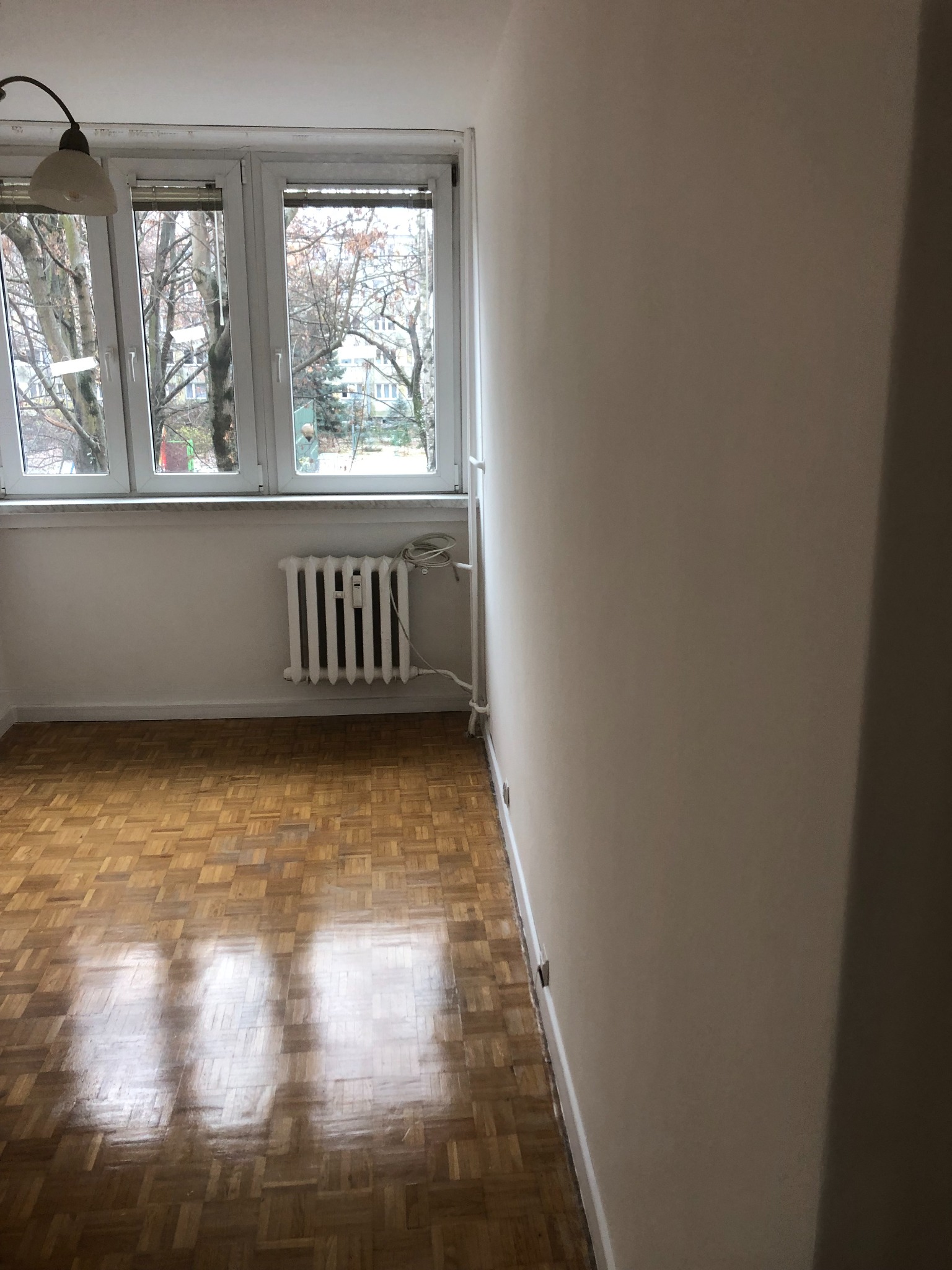 Pusty pokój z parkietem, białymi ścianami i grzejnikiem pod oknem. Widok na drzewa bez liści za oknem.