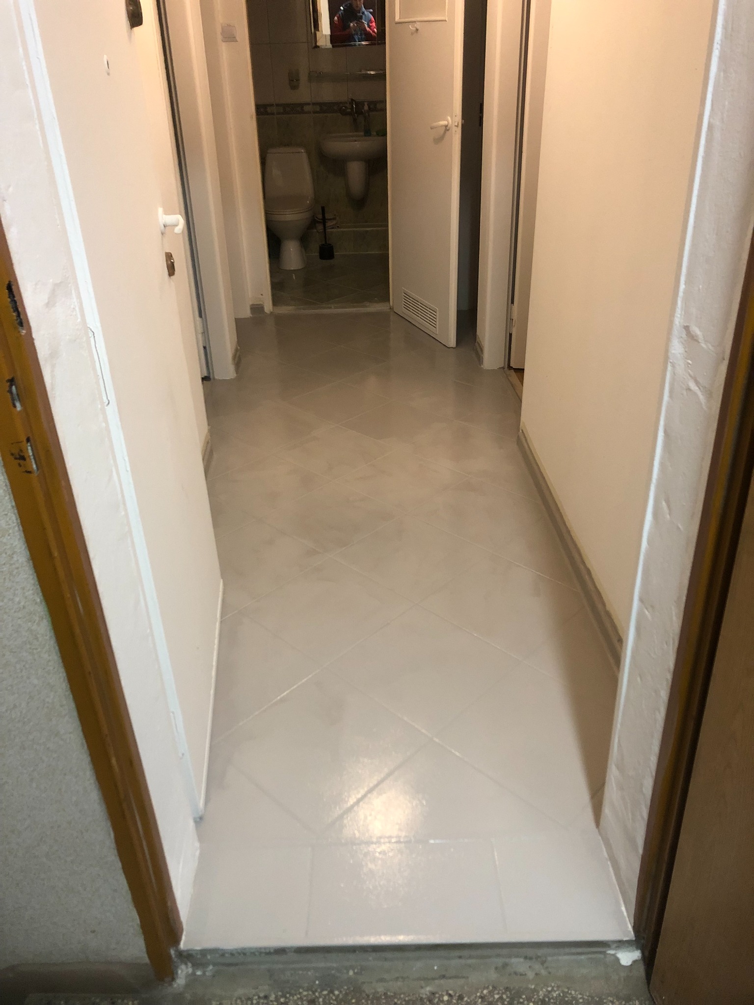 Świeżo pomalowany korytarz z jasnymi płytkami podłogowymi, widok na otwarte drzwi do łazienki w tle.