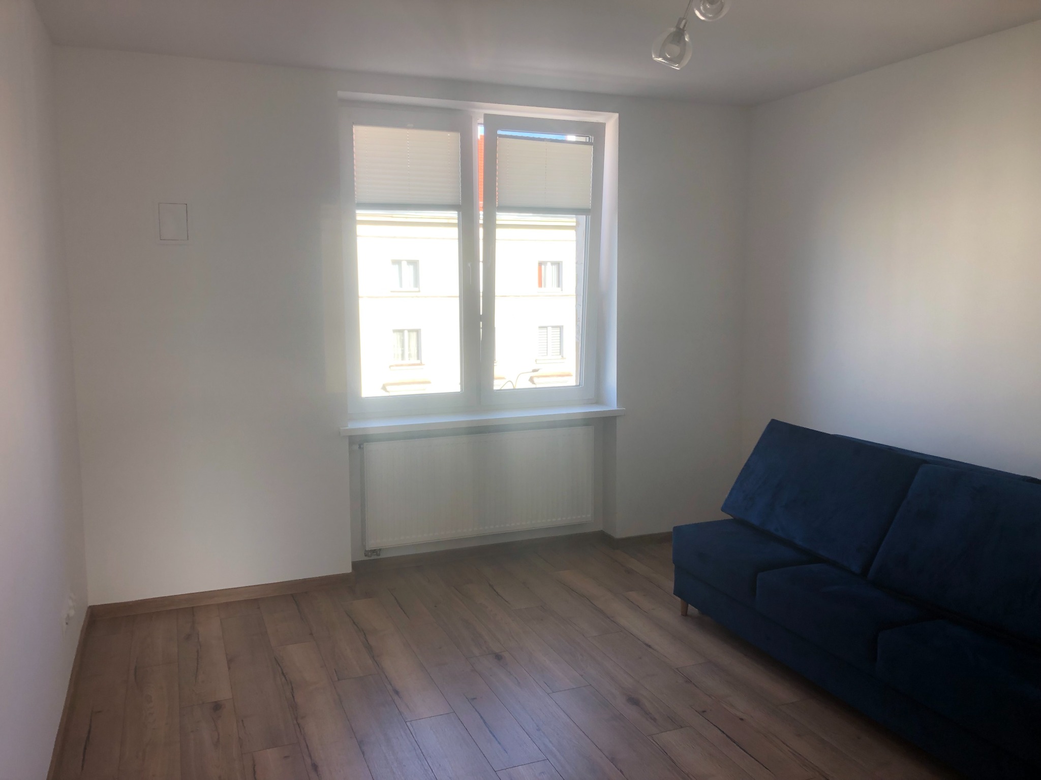 Jasne, puste pomieszczenie z drewnianą podłogą, białymi ścianami, oknem z żaluzjami i granatową sofą w rogu.