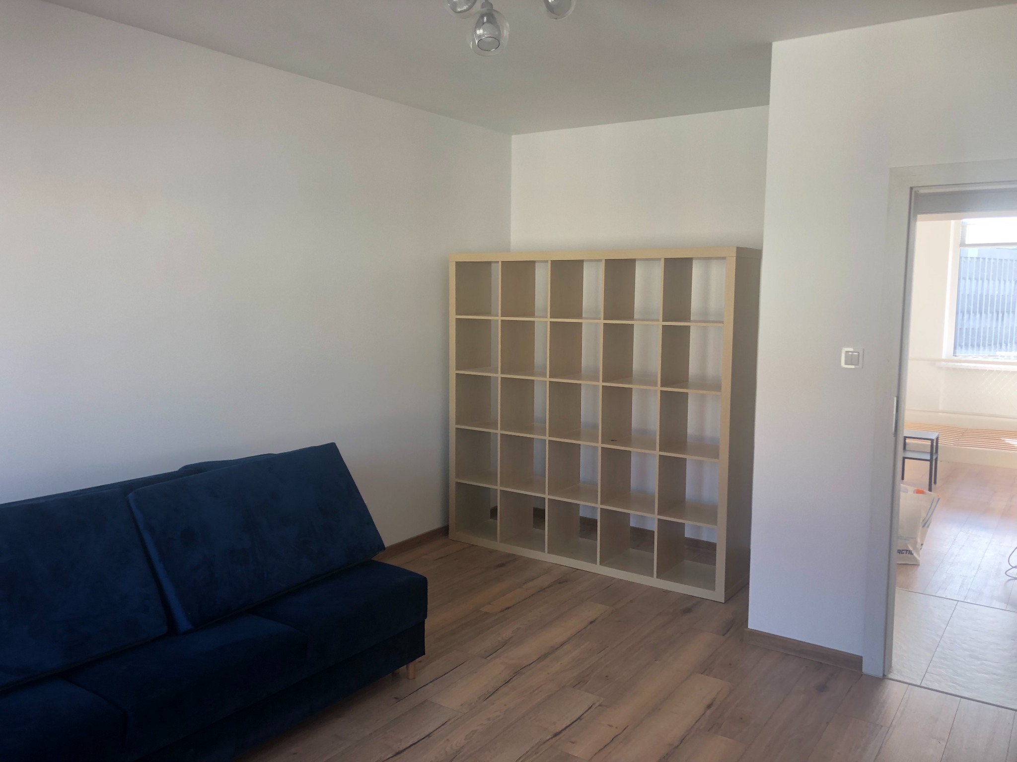 Pusty pokój z ciemnoniebieską sofą, jasną drewnianą podłogą i regałem z kwadratowymi przegródkami, świeżo po remoncie, z widocznymi białymi ścianami.