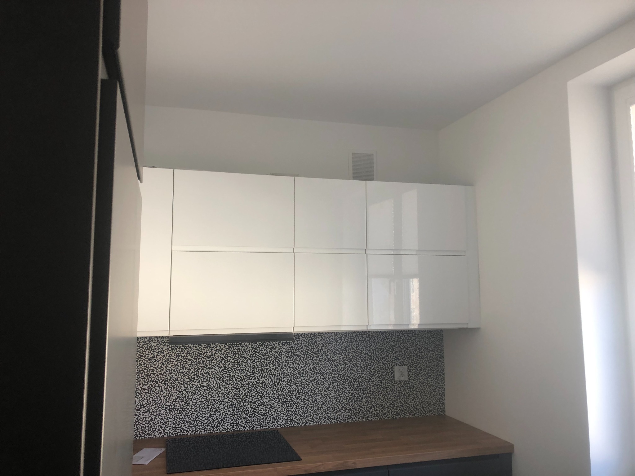 Nowoczesna kuchnia z białymi szafkami wiszącymi, mozaikową ścianą nad blatem i drewnianym blatem, widziana z dołu, z fragmentem czarnej szafki po lewej stronie.