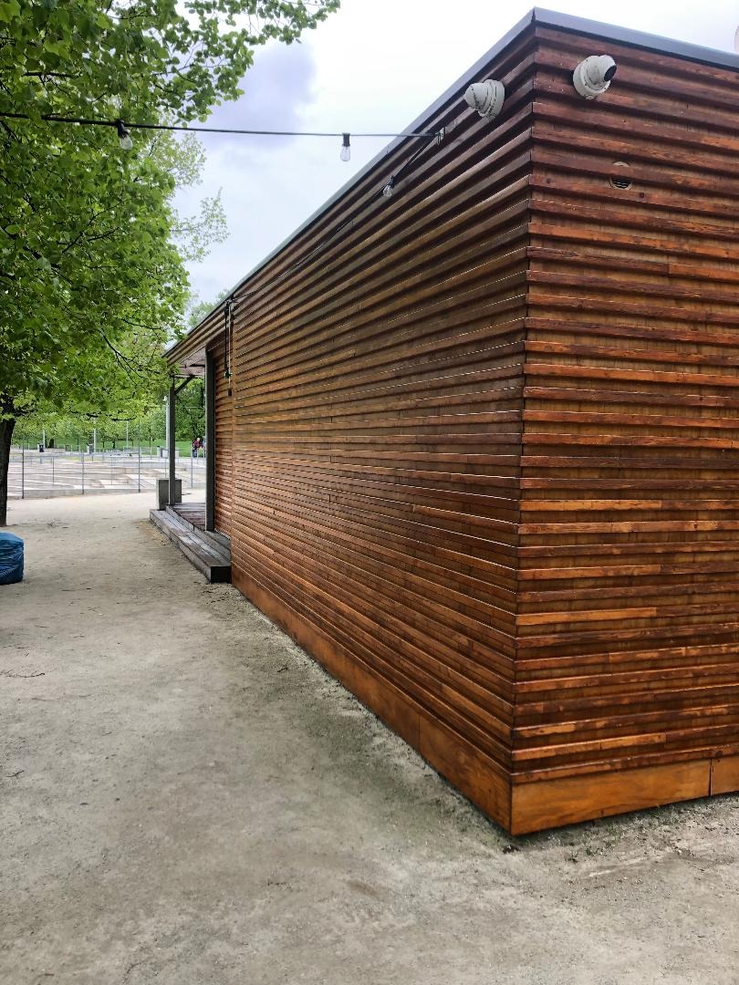 Nowoczesna elewacja budynku obłożona poziomymi deskami z ciemnego drewna, widok z zewnątrz, oświetlenie zewnętrzne zamontowane na górnej krawędzi, z boku widoczne drzewa i chodnik.