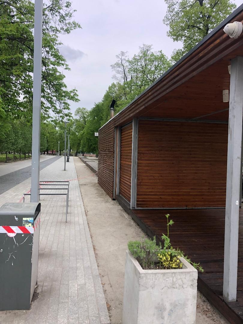 Drewniana altana z elementami metalowymi w parku miejskim, widoczna z boku, obok ścieżki spacerowej z koszem na śmieci i betonową donicą z roślinami.