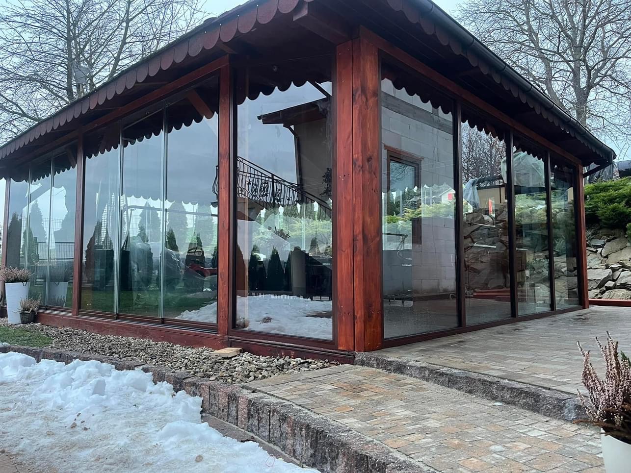 Zadaszona konstrukcja z przeszklonymi ścianami na posesji, widoczny fragment chodnika z kostki brukowej i resztki śniegu.