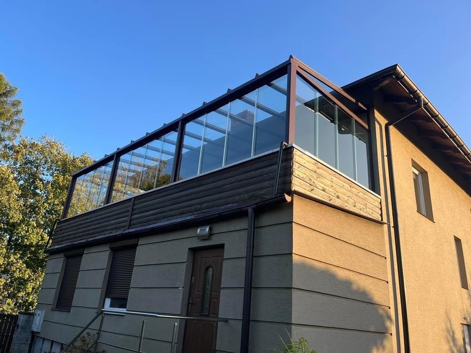 Metalowa konstrukcja z przeszkleniami na balkonie domu jednorodzinnego, widok z dołu pod kątem w słoneczny dzień.