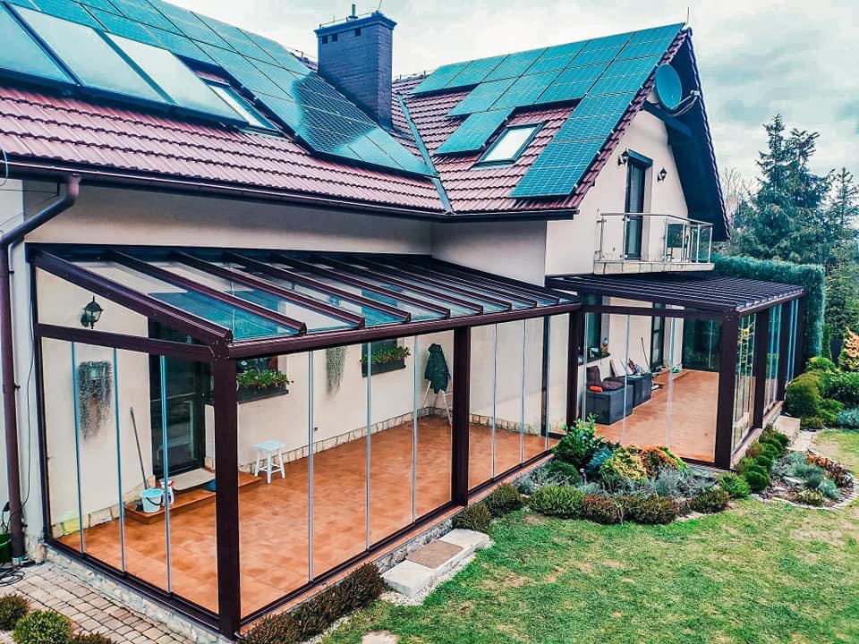 Metalowa pergola z przeszklonymi ścianami przylegająca do domu z panelami słonecznymi na dachu, widok z ogrodu.