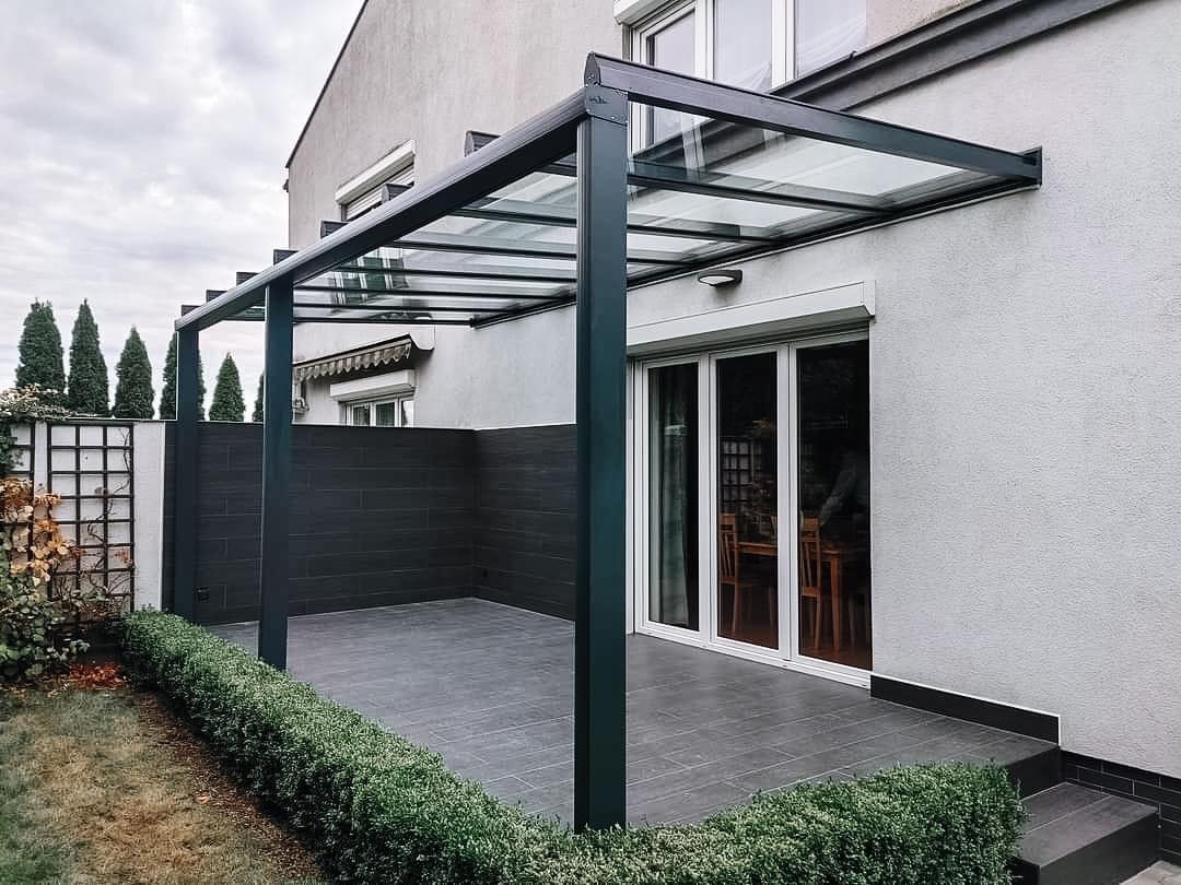 Nowoczesna, czarna konstrukcja z przeszklonym dachem, przylegająca do jasnego budynku z dużymi oknami, otoczona niskim żywopłotem i ciemnymi płytkami tarasowymi.