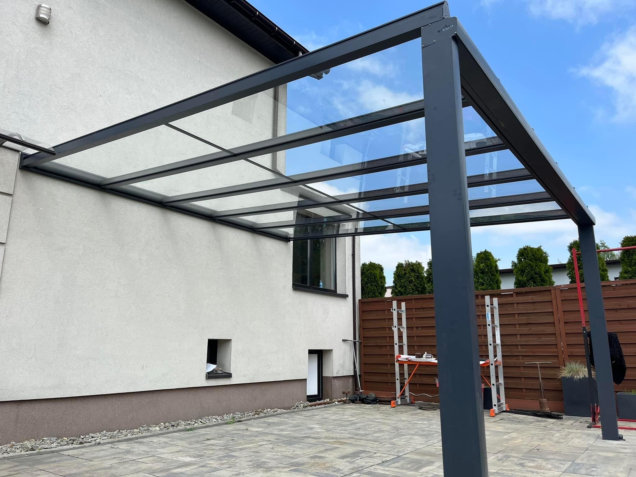 Nowoczesna konstrukcja z metalu i szkła, przylegająca do ściany domu, z widocznym drabiniastym rusztowaniem i narzędziami sugerującymi niedawny montaż.