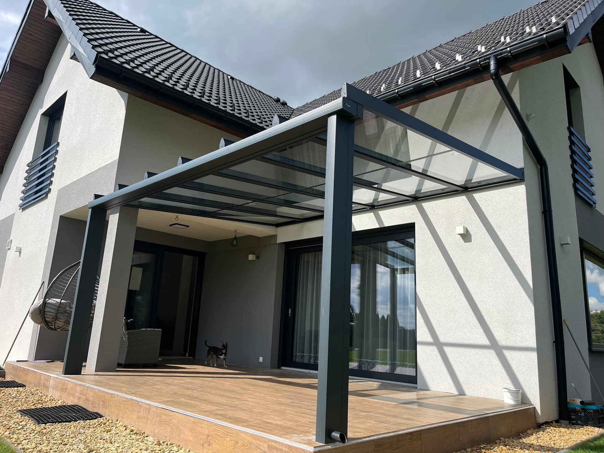 Nowoczesna, szara konstrukcja z przeszklonym dachem na drewnianym tarasie w Mikołowie, przylegająca do jasnego domu z ciemną dachówką. W tle widoczny mały pies.