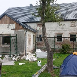 Maciej Malek - Dom w trakcie ocieplania elewacji: widoczne rusztowania, pracownik na wysokościach, częściowo ocieplona ściana, porozrzucane fragmenty styropianu i niebieski namiot na trawniku.