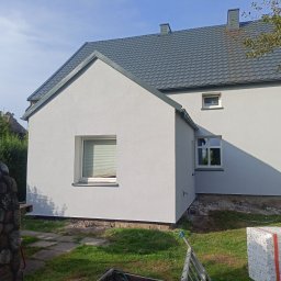 Maciej Malek - Świeżo pomalowana, jasnoszara elewacja domu z szarą dachówką, widok z boku, z widocznymi fragmentami ogrodu i stosem styropianu przy ścianie.