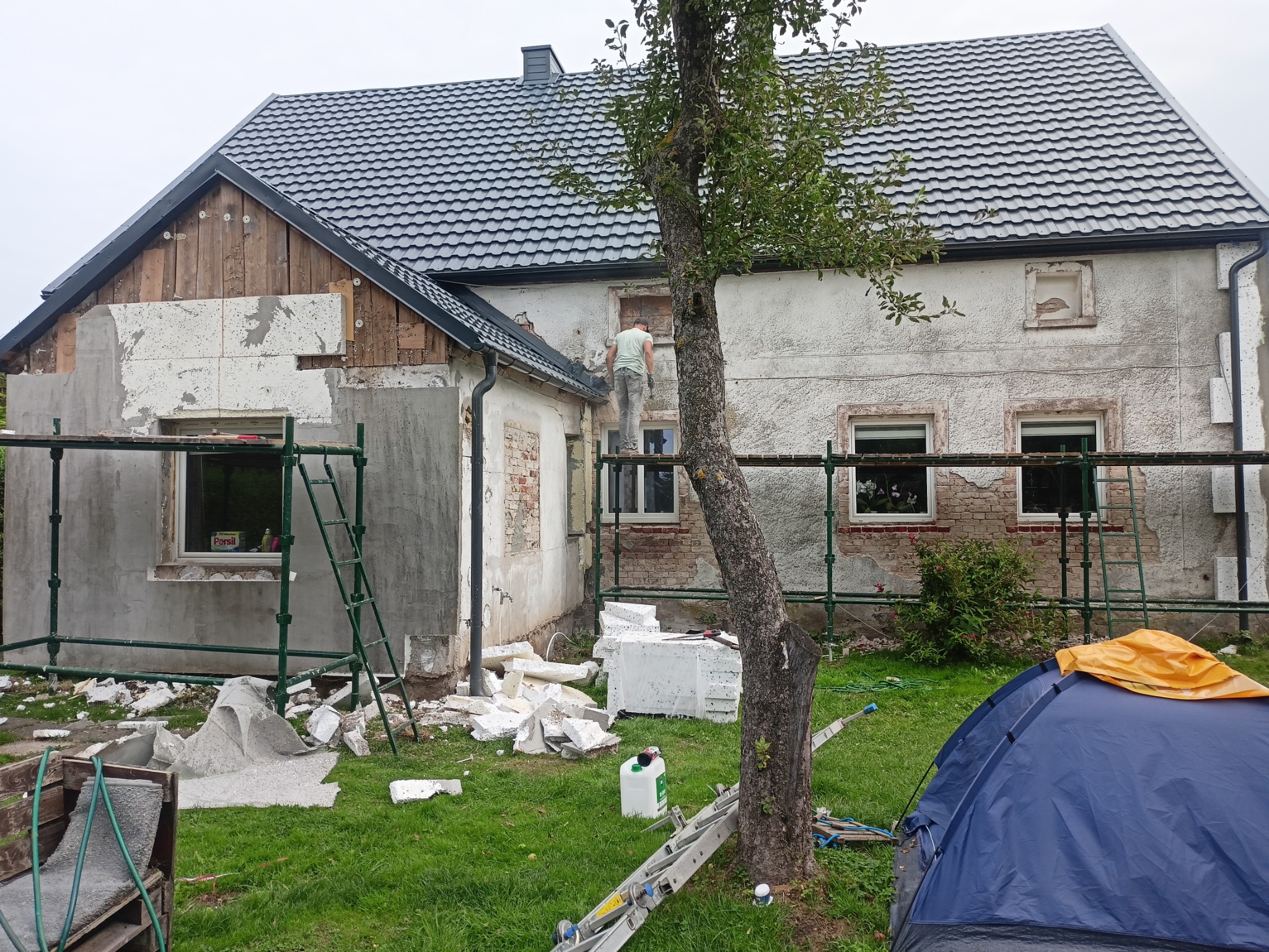 Dom w trakcie ocieplania elewacji: widoczne rusztowania, pracownik na wysokościach, częściowo ocieplona ściana, porozrzucane fragmenty styropianu i niebieski namiot na trawniku.
