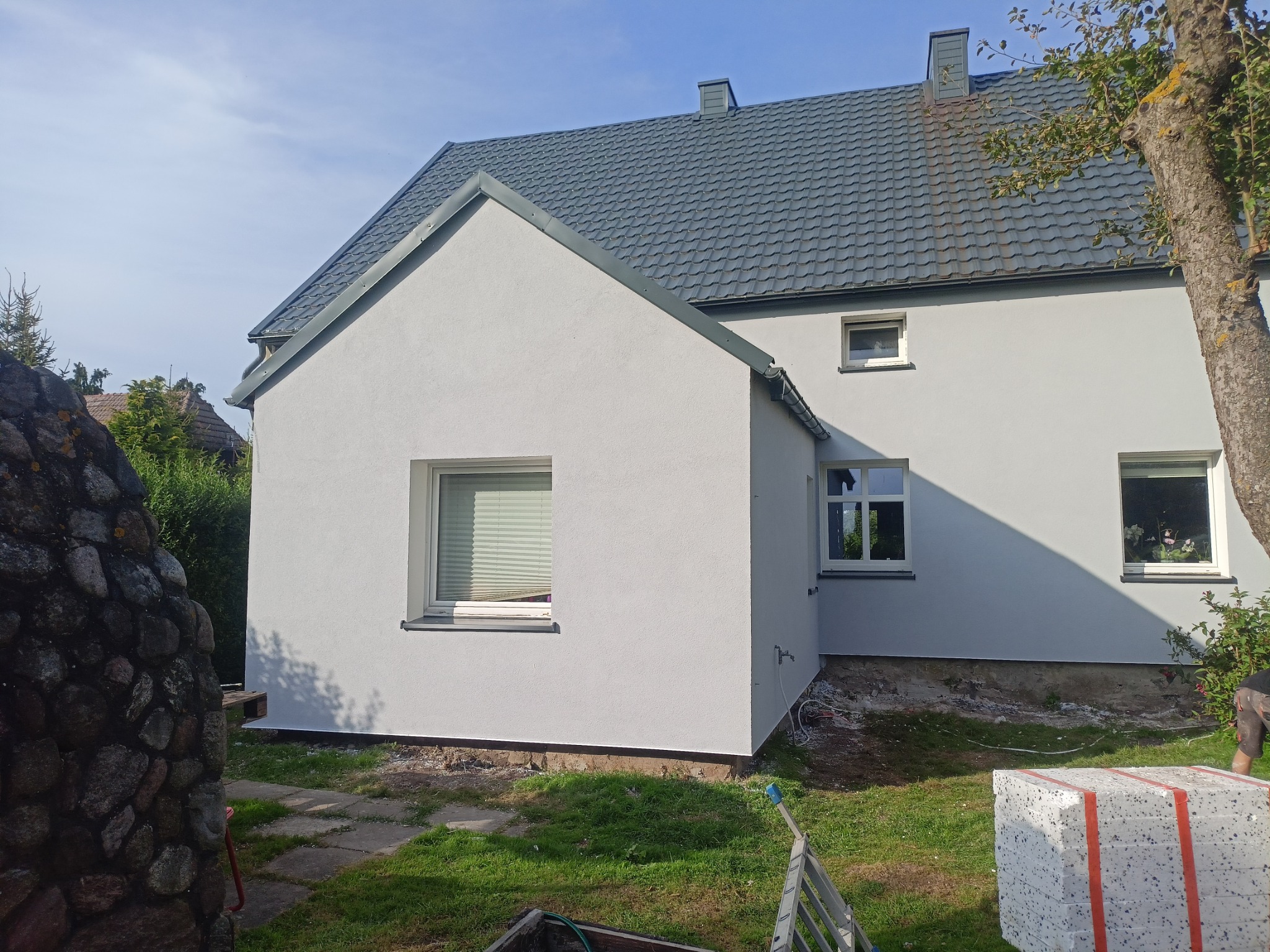 Świeżo pomalowana, jasnoszara elewacja domu z szarą dachówką, widok z boku, z widocznymi fragmentami ogrodu i stosem styropianu przy ścianie.