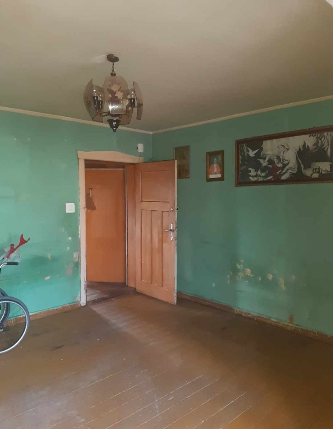 Pokój z zielonymi ścianami przed remontem, widoczne ślady po usuniętych tapetach, stara drewniana podłoga, otwarte drzwi i rower oparty o ścianę.