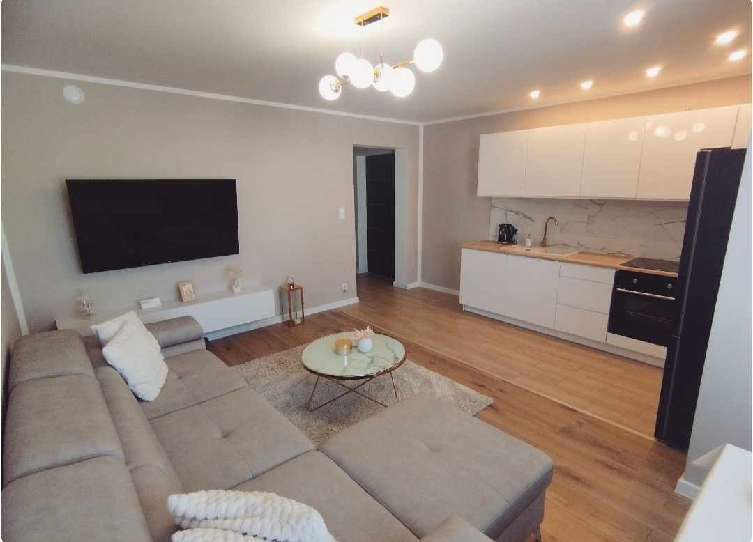 Jasne, nowocześnie urządzone wnętrze salonu z aneksem kuchennym, beżowa sofa z poduszkami, okrągły stolik kawowy na dywanie, białe meble kuchenne z marmurowym backsplash'em i drewnianym blatem...