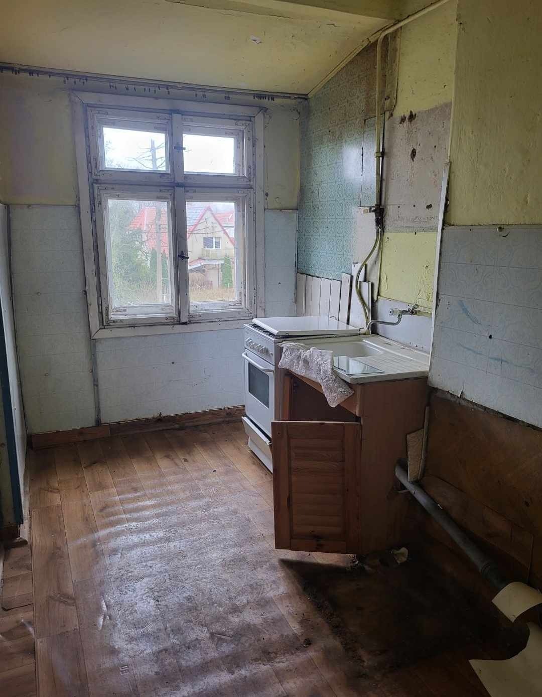 Zniszczona kuchnia z odsłoniętymi fragmentami ścian, starą kuchenką i szafką z otwartymi drzwiczkami, widok z okna na dom z czerwoną dachówką.