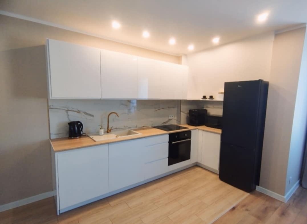 Nowoczesna kuchnia z białymi szafkami, drewnianym blatem i marmurowym backsplashiem, uzupełniona złotą armaturą i czarną lodówką.