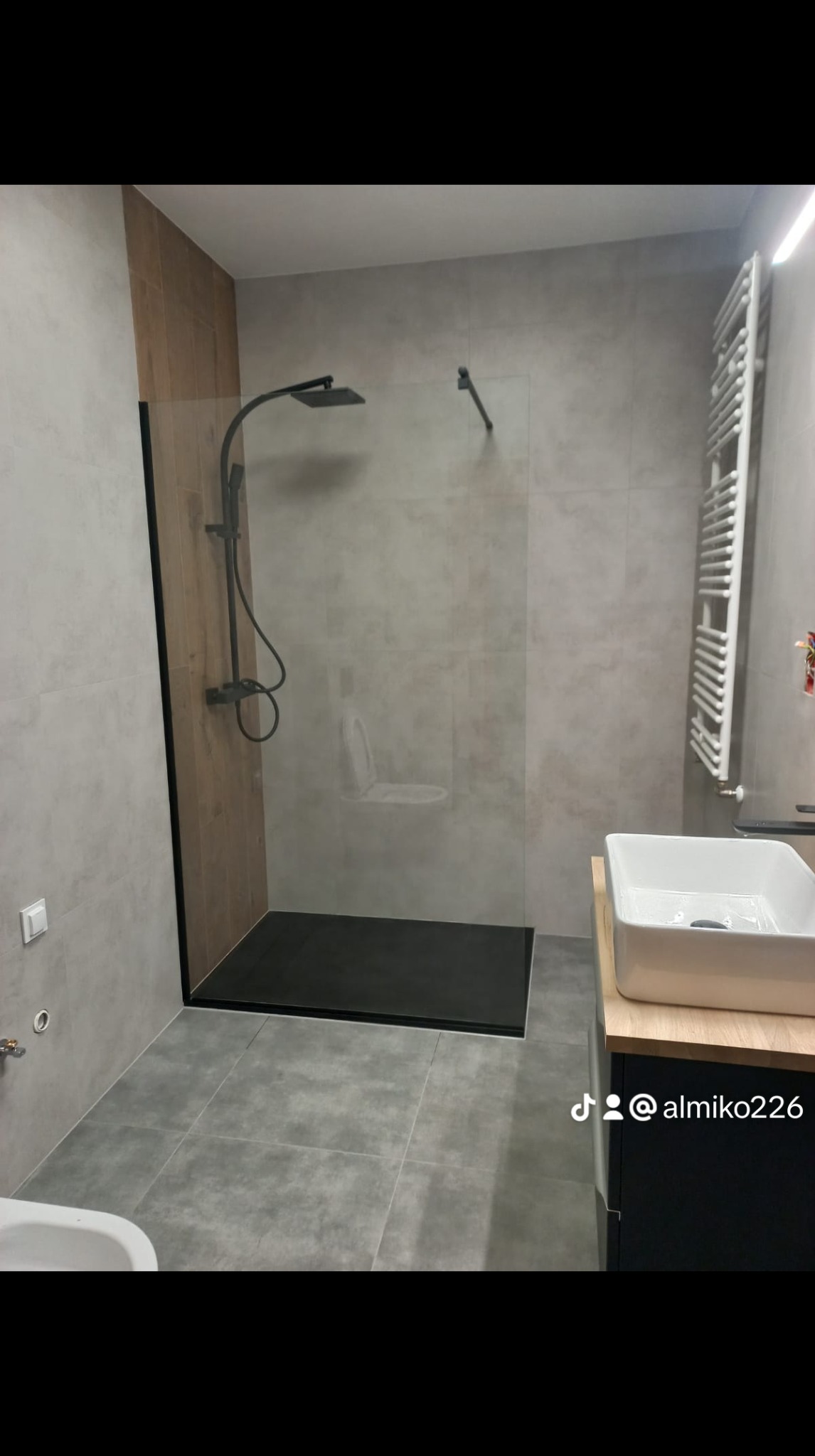 Nowoczesna łazienka z kabiną typu walk-in, czarną armaturą i grzejnikiem drabinkowym, wykończona szarą płytką imitującą beton.