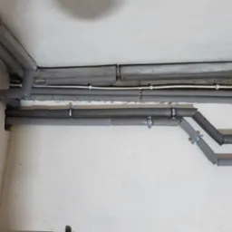 Szare, zaizolowane rury hydrauliczne przymocowane metalowymi uchwytami do ściany i sufitu w surowym wnętrzu. Widoczne łączenia rur i izolacji.