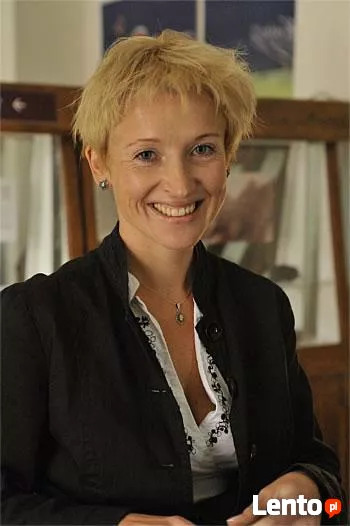 Portret uśmiechniętej kobiety z krótkimi blond włosami, w czarnej marynarce i białej bluzce z delikatnym wzorem, na tle częściowo widocznej witryny ekspozycyjnej.