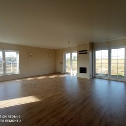 USŁUGI REMONTOWO-WYKOŃCZENIOWE Tomasz Florkowski - Wnętrze przestronnego pokoju po remoncie, z parkietem ułożonym w jodełkę, jasnymi ścianami, dużymi oknami i minimalistycznym kominkiem.