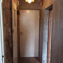 USŁUGI REMONTOWO-WYKOŃCZENIOWE Tomasz Florkowski - Korytarz w trakcie remontu, widoczne odsłonięte cegły, surowy beton, drewniana podłoga i wisząca lampa z abażurem, drzwi wejściowe białe.