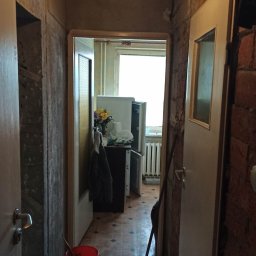 USŁUGI REMONTOWO-WYKOŃCZENIOWE Tomasz Florkowski - Korytarz w trakcie remontu: odsłonięta cegła, surowe ściany, widoczny fragment kuchni z kwiatami i lodówką, podłoga częściowo zakryta, czerwone wiadro i miotła w korytarzu, wisząca lampa z żarówką.