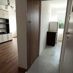 USŁUGI REMONTOWO-WYKOŃCZENIOWE Tomasz Florkowski - Korytarz po remoncie: szare płytki podłogowe, zabudowa szafy z drewnopodobną okleiną, widok na salon z drewnianą podłogą i białe meble.