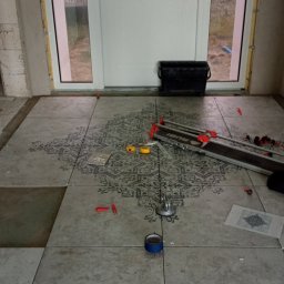 USŁUGI REMONTOWO-WYKOŃCZENIOWE Tomasz Florkowski - Układanie szarych płytek podłogowych z wzorem w korytarzu, widoczna przecinarka do płytek Rubi, poziomica, przyssawka do płytek i rolka niebieskiej taśmy malarskiej.