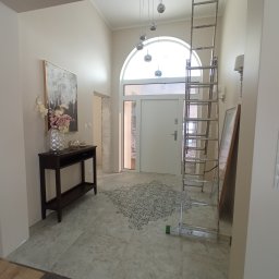 USŁUGI REMONTOWO-WYKOŃCZENIOWE Tomasz Florkowski - Jasne wnętrze w trakcie remontu: widoczna drabina oparta o ścianę, dekoracyjny wzór na podłodze, konsola z kwiatami i obrazem, oraz wejście z łukowym oknem.