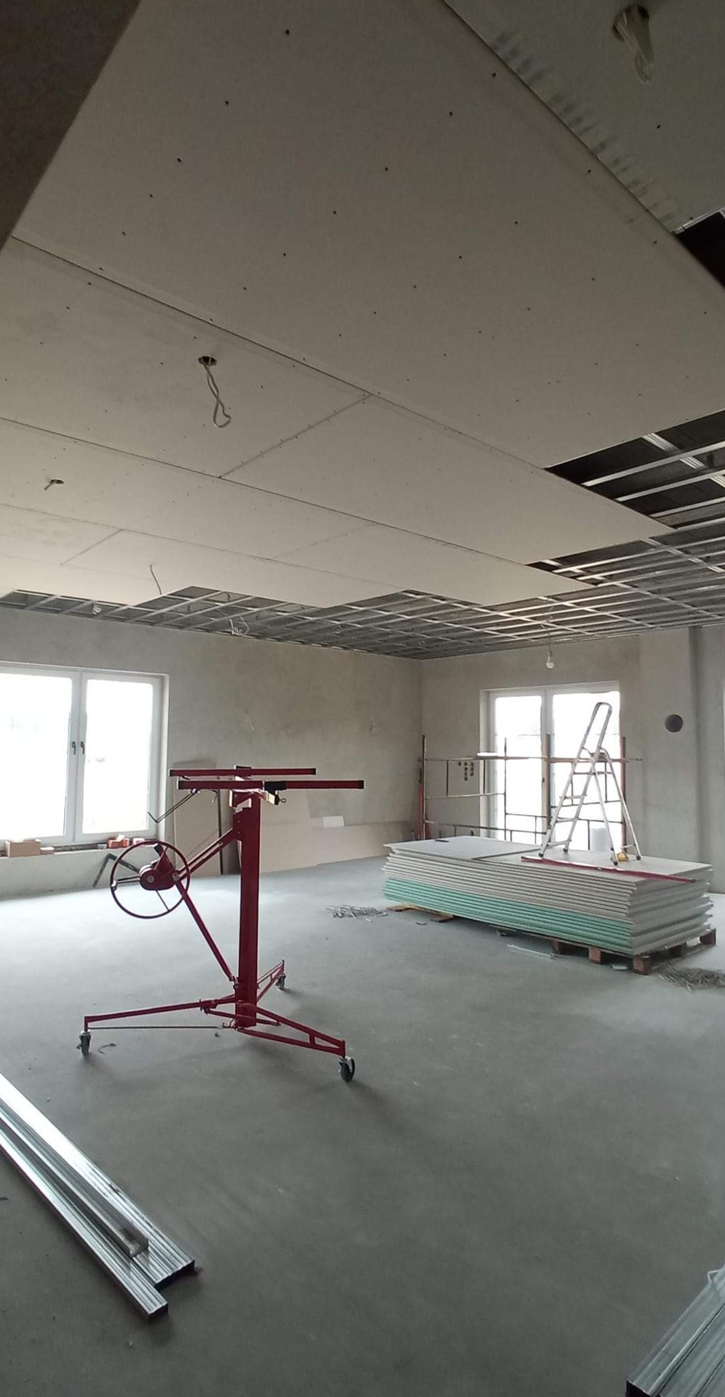 Montaż sufitu podwieszanego z płyt gipsowo-kartonowych w biurze, widoczny stelaż, podnośnik do płyt, ułożone płyty, drabina i okna.