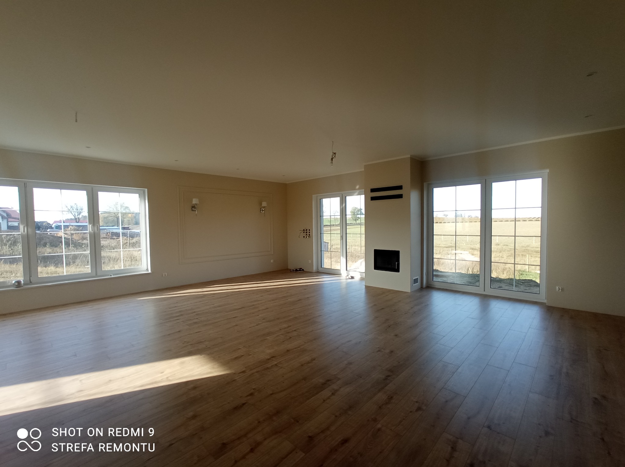 Wnętrze przestronnego pokoju po remoncie, z parkietem ułożonym w jodełkę, jasnymi ścianami, dużymi oknami i minimalistycznym kominkiem.