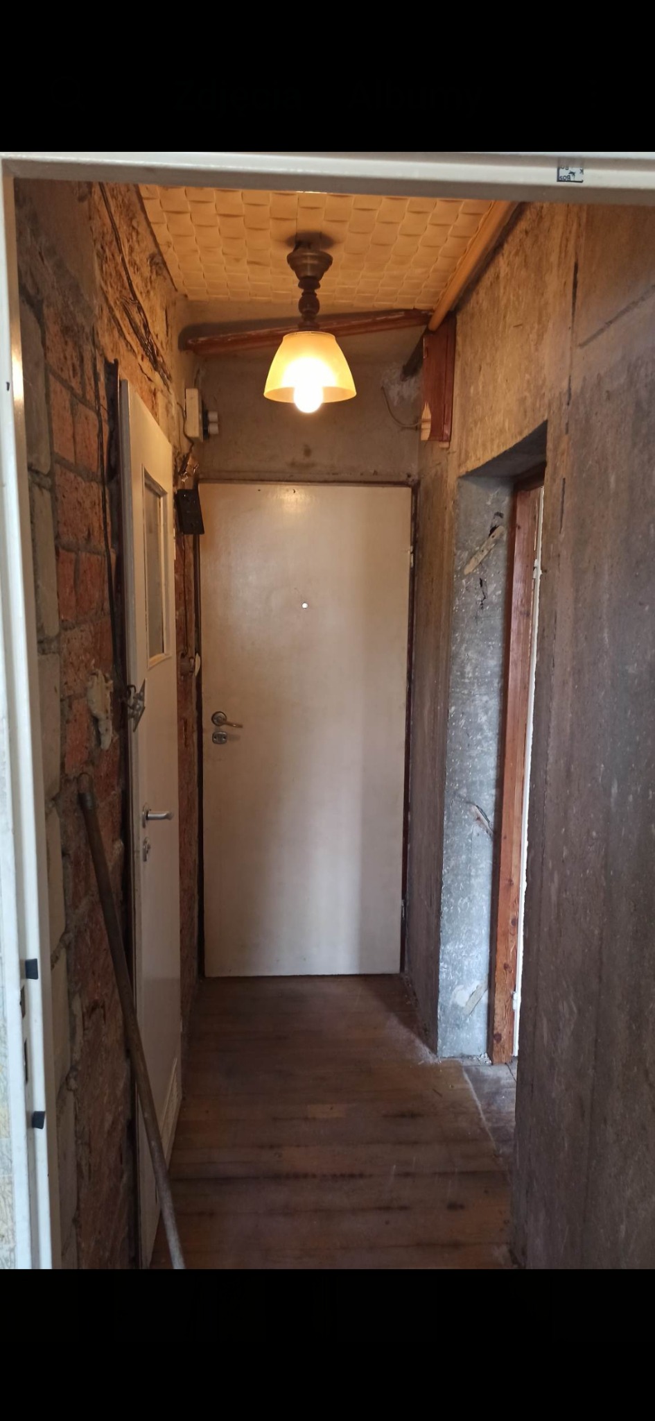 Korytarz w trakcie remontu, widoczne odsłonięte cegły, surowy beton, drewniana podłoga i wisząca lampa z abażurem, drzwi wejściowe białe.