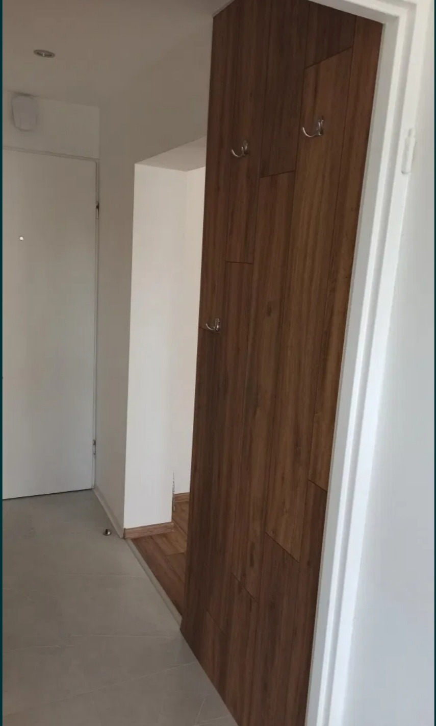 Drewniana zabudowa z haczykami w przedpokoju, imitująca panele ścienne, widok na białe drzwi wejściowe i fragment podłogi w mieszkaniu.