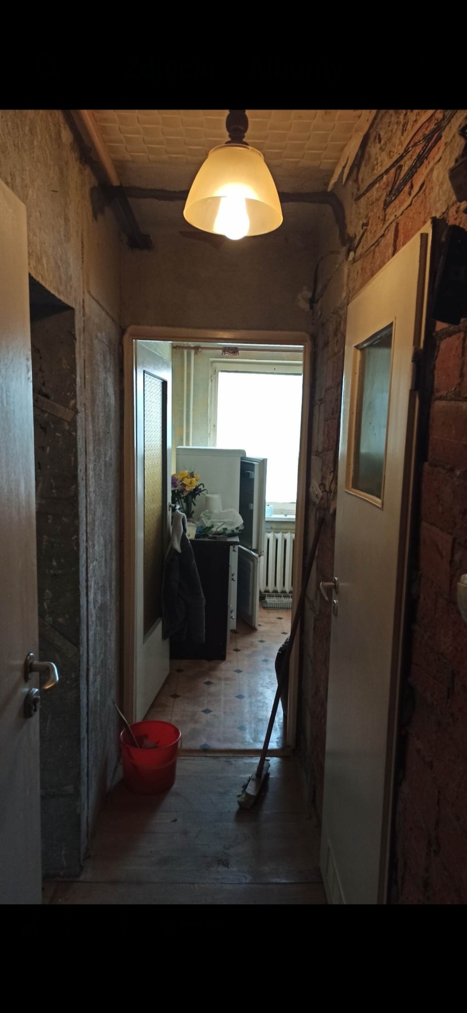 Korytarz w trakcie remontu: odsłonięta cegła, surowe ściany, widoczny fragment kuchni z kwiatami i lodówką, podłoga częściowo zakryta, czerwone wiadro i miotła w korytarzu, wisząca lampa z żarówką.
