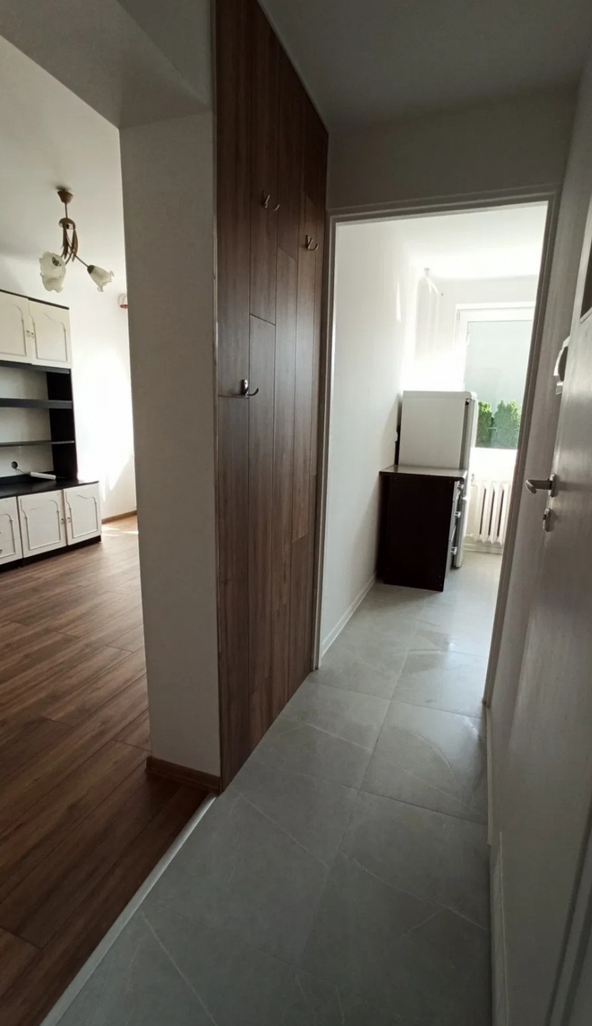 Korytarz po remoncie: szare płytki podłogowe, zabudowa szafy z drewnopodobną okleiną, widok na salon z drewnianą podłogą i białe meble.