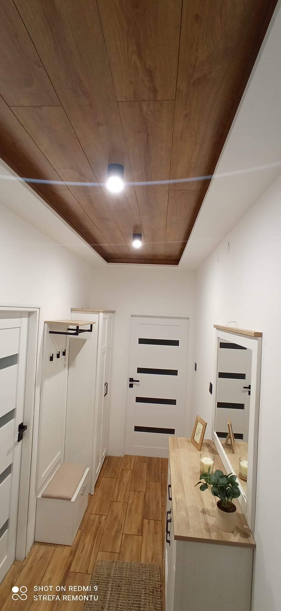Nowoczesny przedpokój z drewnianym sufitem podwieszanym z oświetleniem punktowym, białymi ścianami, panelami podłogowymi, szafą z wieszakiem, komodą z lustrem i dekoracjami.