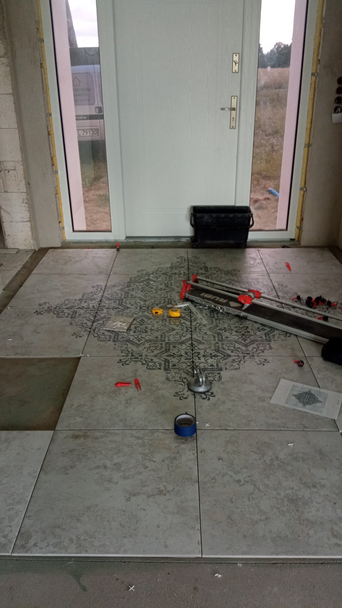 Układanie szarych płytek podłogowych z wzorem w korytarzu, widoczna przecinarka do płytek Rubi, poziomica, przyssawka do płytek i rolka niebieskiej taśmy malarskiej.