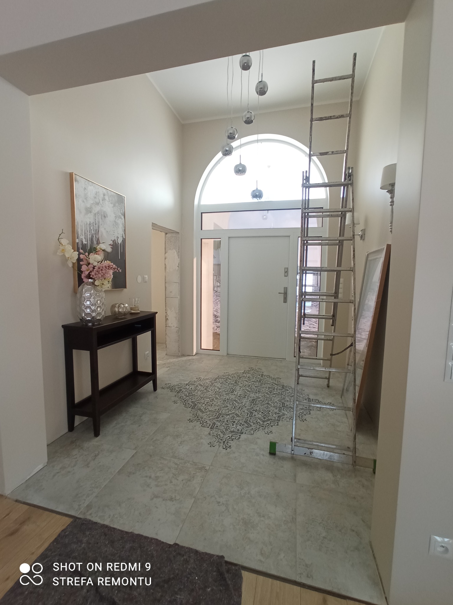 Jasne wnętrze w trakcie remontu: widoczna drabina oparta o ścianę, dekoracyjny wzór na podłodze, konsola z kwiatami i obrazem, oraz wejście z łukowym oknem.