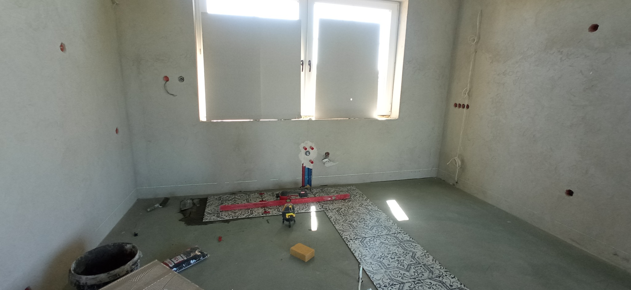 Wnętrze w trakcie układania płytek podłogowych z wzorem ornamentu, widoczne narzędzia glazurnicze, poziomica laserowa i puste puszki elektryczne w ścianie.