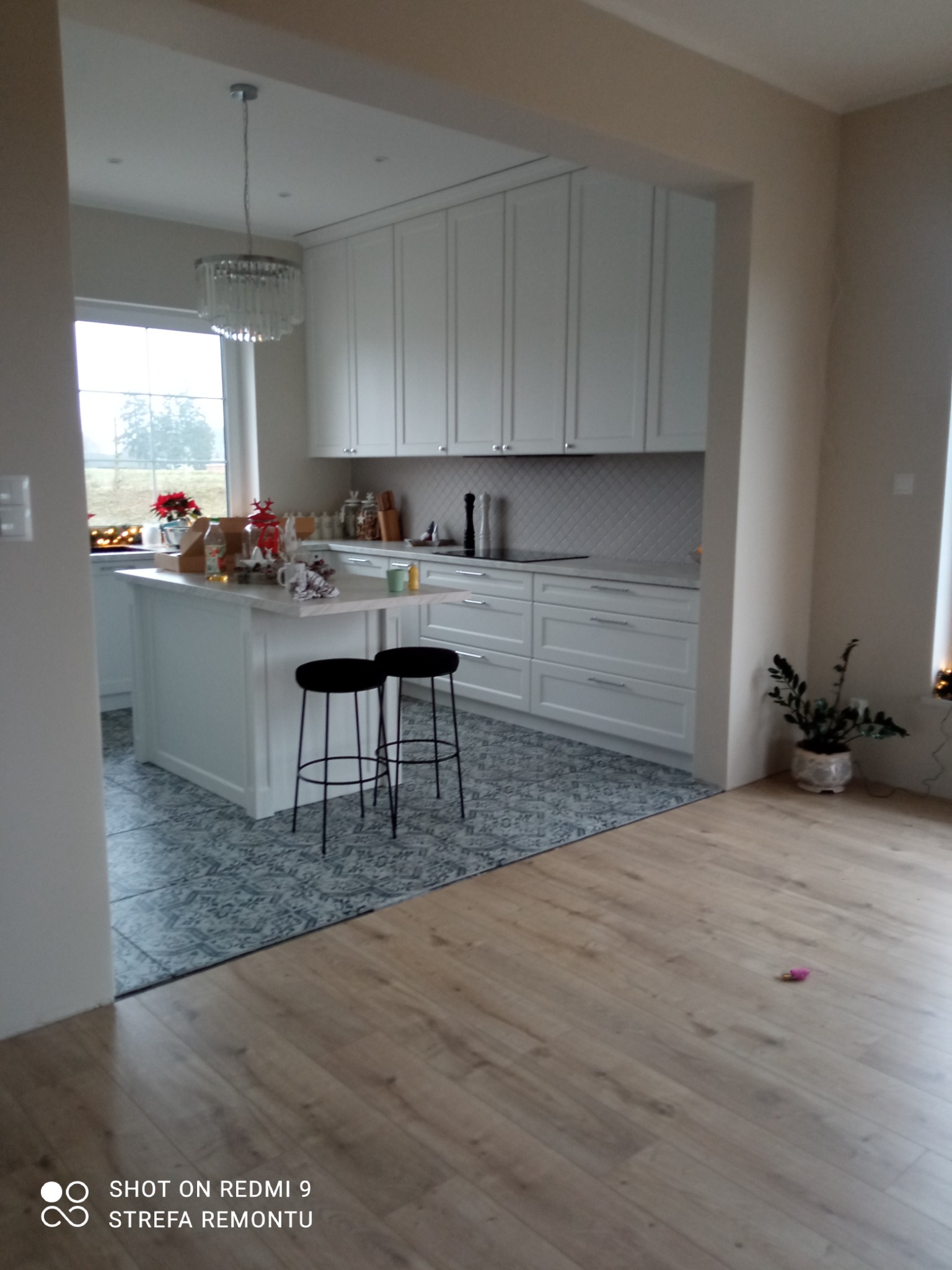 Jasna, przestronna kuchnia z wyspą i dwoma czarnymi hokerami, widok z salonu na wykończoną kuchnię z białymi szafkami i wzorzystą podłogą, dekoracje świąteczne na wyspie kuchennej.