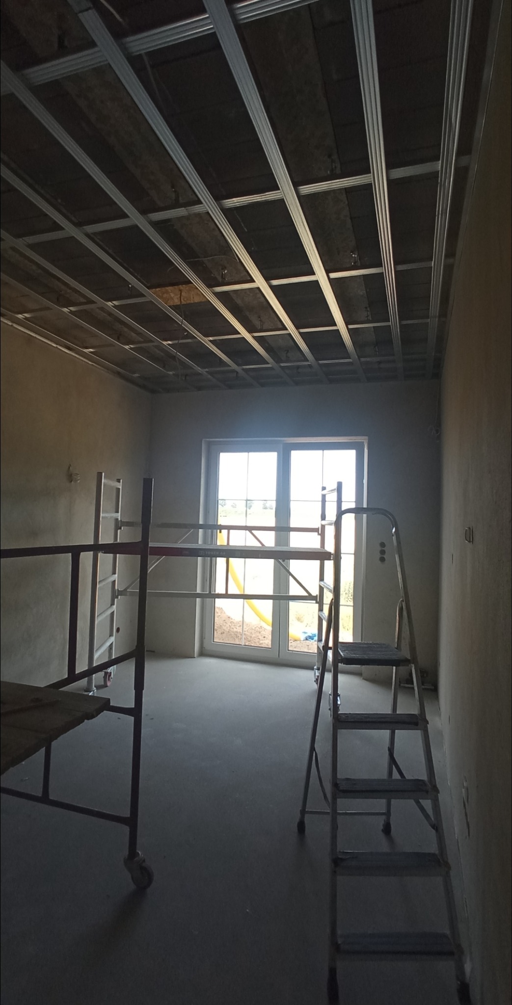 Metalowa konstrukcja stelaża podwieszanego sufitu z widocznymi elementami izolacji, rusztowanie i drabina w surowym wnętrzu z oknem.