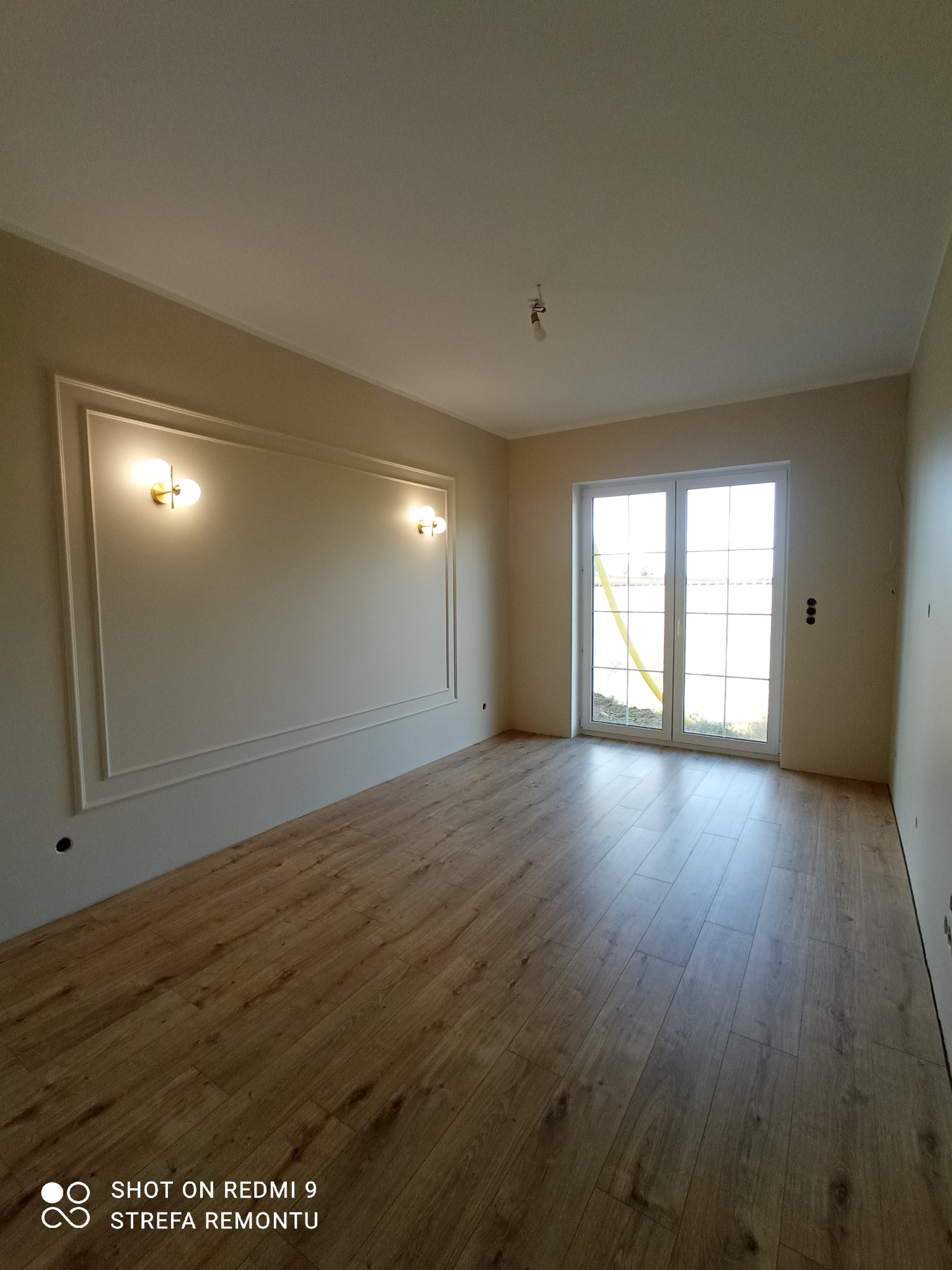 Pusty pokój z panelami podłogowymi w odcieniach drewna, kremowe ściany z ozdobną ramą i kinkietami, białe okno balkonowe z widokiem na zewnątrz.