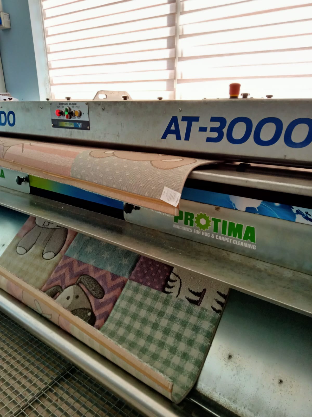 Automatyczna maszyna AT-3000 marki PROTIMA w trakcie czyszczenia dywanów, z widocznymi dywanami z dziecięcymi wzorami w środku.