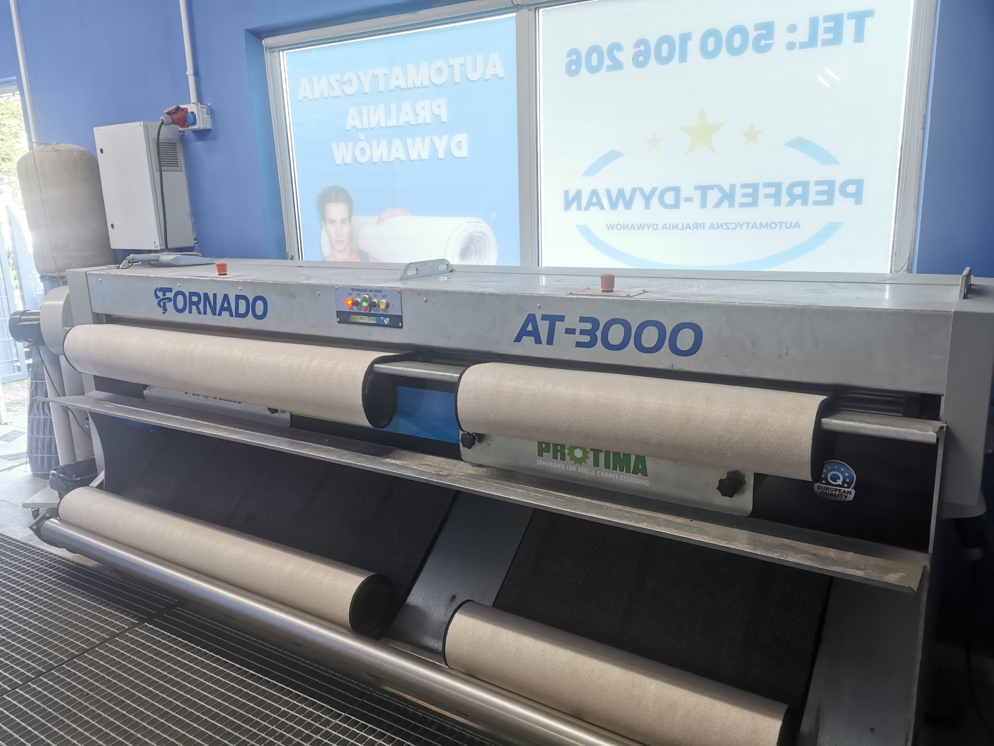 Profesjonalna maszyna do czyszczenia dywanów marki Tornado AT-3000 z wałkami i systemem transportu dywanów, widoczny napis PROTIMA, w tle okno z reklamą firmy.