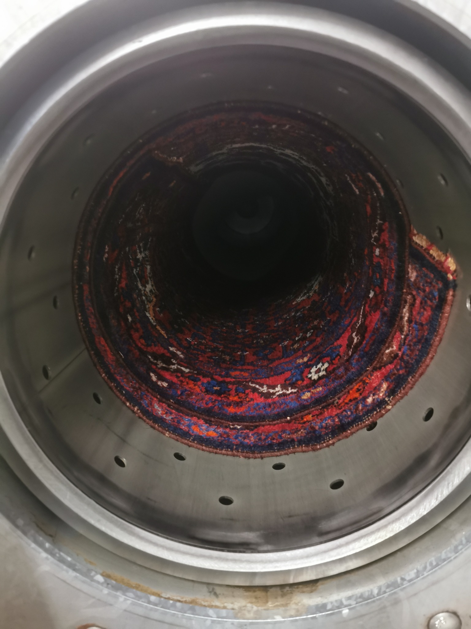 Wnętrze przemysłowej pralki z orientalnym dywanem zwiniętym w spiralę, widoczne otwory wentylacyjne w metalowej obudowie.